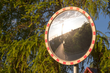 Lustrzany znak drogowy ( u-18) na tle iglastych drzew . W lustrze odbija się wizerunek domów i ulic oraz zachodzące słońce . Znak na peryferiach - Ostrowiec Świetokrzyski - miasta .