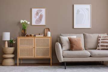 Interior design of cozy and warm living room with mock up poster frame, rattan sideboard, beige sofa, brown pillow, vase with dried flowers, sculpture and personal accessories. Home decor. Template.