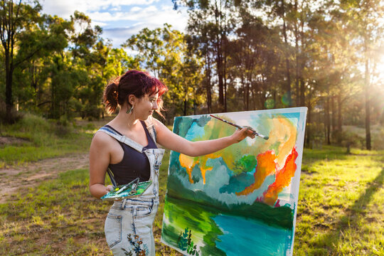 Backlit young artist in forest clearing working on large landscape painting