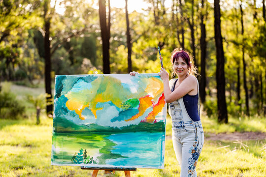 Smiling portrait of happy young woman artist in forest clearing with beginning of painting