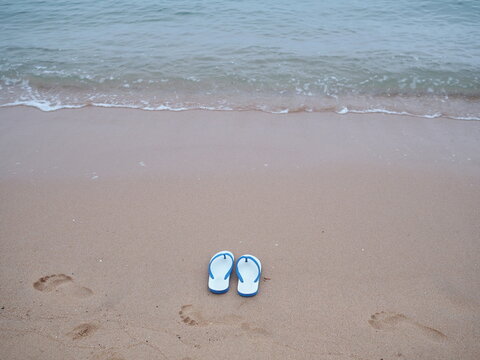 Pair of white  Flip-flops shoes with blue straps rests on sandy beach with small wave in front of the sea. Sponge shoes were removed and left in wet sand,there were blurry images of human footprints 
