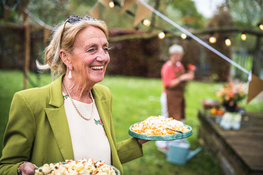 Smiling beautiful senior woman bringing food for party, preparing a lunch and an outdoor party in the garden, healthy and happy elderly people concept