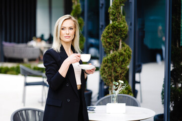 Attractive stylish woman dressed in black elegant suit standing next to the table in cafe drinking cup of cappuccino