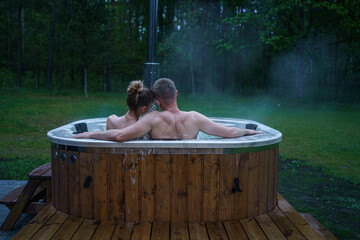 Young happy couple relax in outside rustic hot tub spa surrounded by calm forest scenery