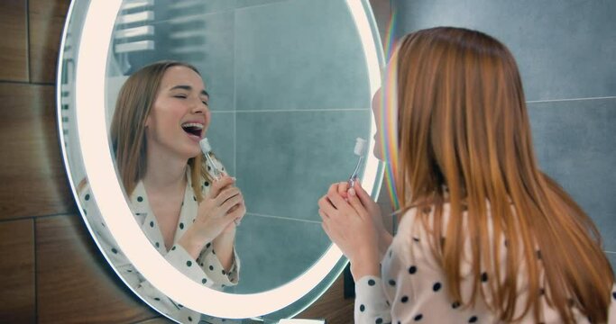 Beautiful caucasian girl brushing teeth in front mirror at bathroom. Healthy teeth, daily hygiene concept.