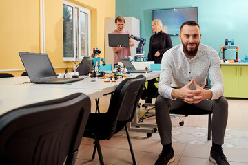 A man sitting in a robotics laboratory while his colleagues in the background test new, cutting edge robotic inventions.