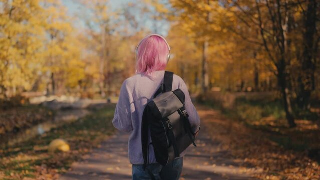 Cheerful pink-haired woman in headphones moves to music in park, back view