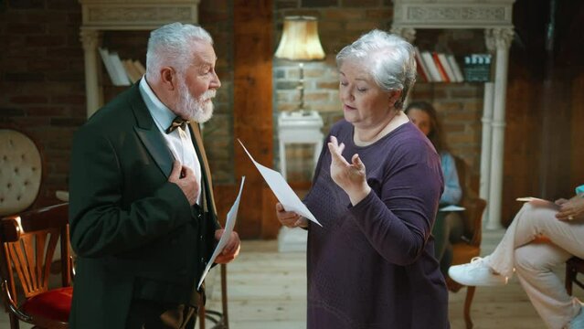 Medium shot of an old actor and elderly actress rehearsing a scene in the theater while holding their scripts