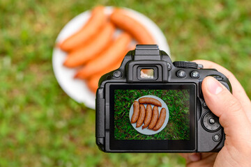 Kiełbasa grillowa na ekranie aparatu fotograficznego i leżąca na talerzu na trawie na świeżym powietrzu  - obrazy, fototapety, plakaty
