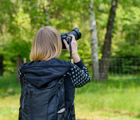 Kobieta w blond włosach z plecakiem na plecach fototrafująca obiekt w lesie, lato i fotografka - obrazy, fototapety, plakaty