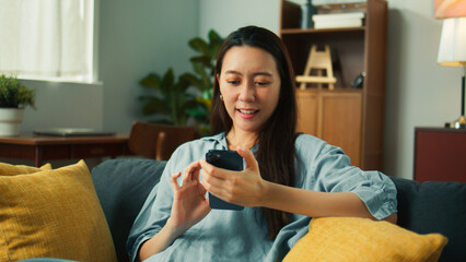Happy beautiful asian woman sit on couch holding mobile phone in hands chatting and playing social...