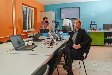 A woman sitting in a laboratory and solving problems and analyzing the robot's verification. In the background, colleagues are talking at an online meeting