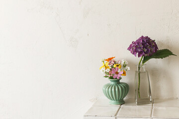 Stylish purple hydrangea and wildflowers bouquet on tile shelf on rustic wall background. Beautiful summer flowers in vase gathered from garden, floral arrangement in modern room in home