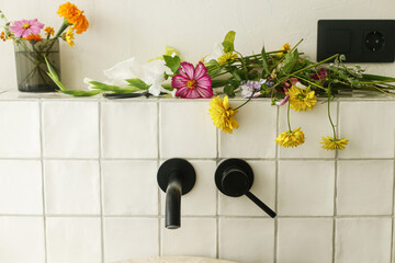 Beautiful flowers in modern bathroom with stylish sink, black faucet and white tiles. Summer flowers arrangement gathered from garden in modern room in home