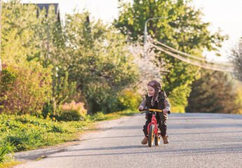 Fototapeta premium A cheerful little boy rides a bicycle outdoors. A happy child walks in the spring park. The baby is dressed in a fashionable vest, overalls. An aviator's hat is on his head.