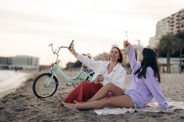 Two happy young women with bike on the beach, making a toast on video call with online friends, raising bottle of beer and smiling, chilling, relaxing, spending time at sea shore.