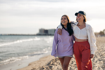 Two happy girlfriends walking with arms around each other on beach. Smiling young women tourists spending time on the beach.