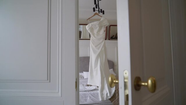 A beautiful white wedding dress hangs on a hanger in the bedroom. Morning of the bride, wedding day. Women's fashionable clothes for the holiday.