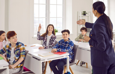 Happy smiling teenager schoolgirl sitting at the desk in the classroom raising her hand up ready to answer to the woman school teacher. Back to school, education, primary school concept.