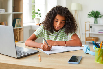 African American teenage girl handwriting in notebook while doing homework using laptop....
