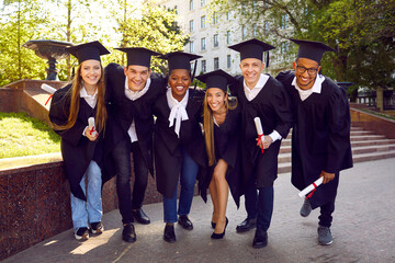Group of multiracial students pose cheerfully for graduation photo outdoors with diplomas. Ready to get success. Celebration of getting new profession in garden near by university. - obrazy, fototapety, plakaty