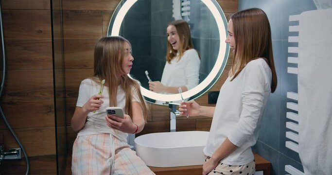 Lifestyle. Beautiful caucasian girls brush their teeth and read messages on phone from bathroom at home. Two girls brushing their teeth and use smartphone, checking social networks