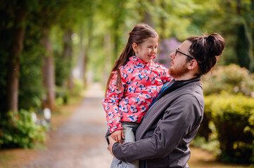 Young long hair bearded man in glasses standing with junior girl holding on hands on road in park