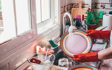 Elderly woman washing crockery in front to the window, wearing red gloves. Home kitchen and colorful dishes under flush of water