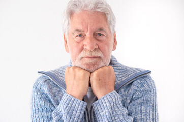 Serious elderly senior man in blue casual sweater looking at camera with hands under chin, isolated on white background