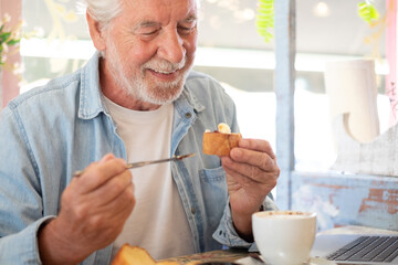 Adult senior man in coffee shop having breakfast with cappuccino and sweet food. Smiling elderly people enjoying retirement and free lifestyle