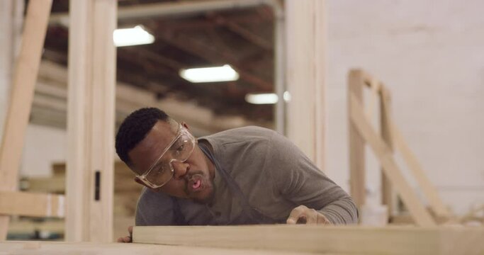 Carpentry, sanding and a man blowing sawdust off of a plank in his factory for professional woodwork. Safety goggles, manual labor and design with a male carpenter working in his creative workshop