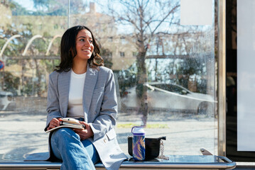 Smiling ethnic woman sitting on bench in city bus stop