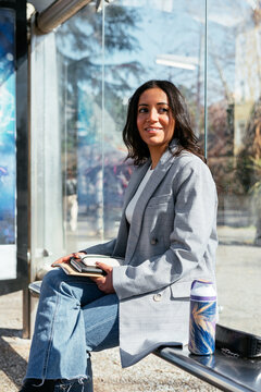 Smiling ethnic woman sitting on bench in city bus stop