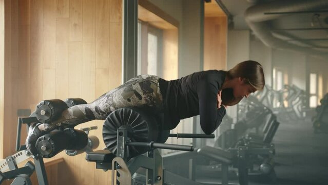 Fit young woman exercises on glute ham developer in fully-equipped gym. Woman horizontally performs hip and back extension with arms crossed on chest. High quality 4k footage