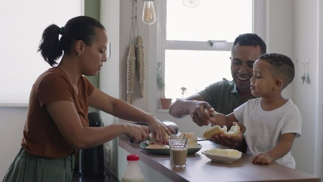 Cheerful mixed race family making breakfast while husband and little son eating and drinking juice at kitchen table
