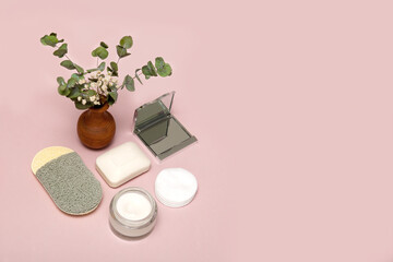 cosmetic accessories face sponge, soap, face cream, mirror, cotton pads on a soft pink background with a vase of greenery and copy space