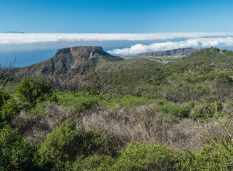 Scenic view on table mountain Fortaleza seen from peak of Alto de at Garajonay mountain. White clouds and El Hiero island above, blue sky. La Gomera, Canary Islands, Spain.