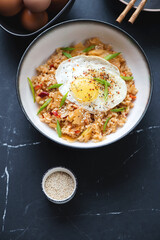 Bowl of kimchi fried rice topped with roasted chicken egg, vertical shot on a black marble background, elevated view