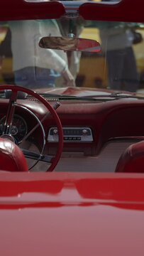 Red leather interior of old retro cabriolet. Old analog radio, rear seat mirror and chairs without headrest. On sunny day, modern muscle car stands in front and people walking.