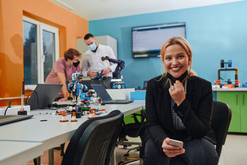 A woman sitting in a laboratory and solving problems and analyzing the robot's verification. In the background, colleagues are talking at an online meeting