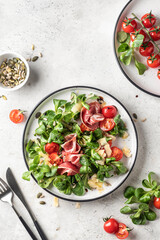 Green salad with ham or prosciutto, corn salad leaves, tomatoes and cheese. Healthy diet lunch on white marble background top view flat lay