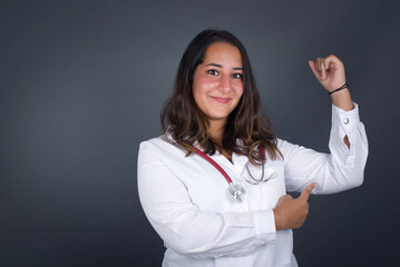 Waist up shot of blonde woman raises hand to show her muscles, feels confident in victory, looks strong and independent, smiles positively at camera, stands against gray background. Sport concept.