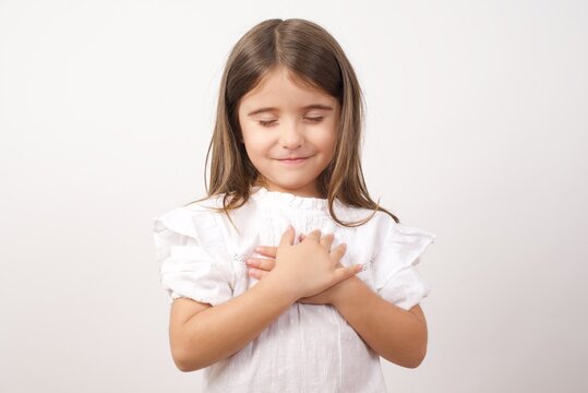 Close up of cute happy small girl isolated on white studio background hold hands at heart chest feel grateful, smiling little child with eyes closed pray thanking god high powers, faith concept