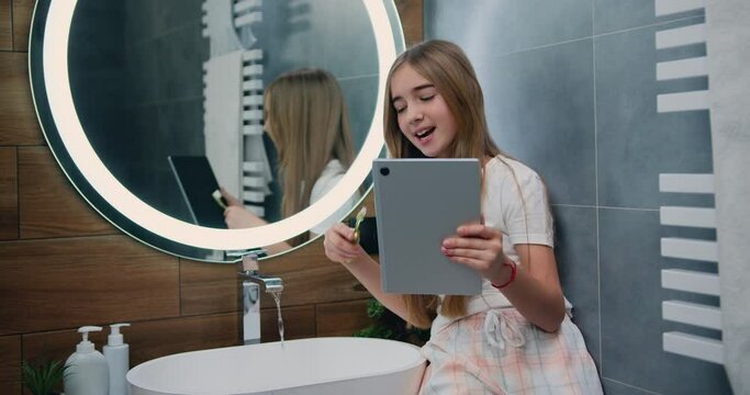 Pretty funny small girl with long hair in pajamas brushing teeth and use tablet device in the morning at bathroom. Cute girl using tablet enjoying cool video or photo content in social network