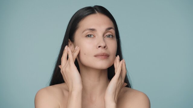 A seminude woman touches her clean and smooth face with wellgroomed fingers. Close up portrait of young woman isolated on blue background. Beauty concept. Natural beauty, cosmetology, skin care.