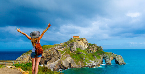 Happy woman with arms raised on the island Gaztelugatxe on the coast of the bay of Biscay- Basque country, near bilbao and Santander- Spain - obrazy, fototapety, plakaty