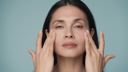 A young woman massages her face with gentle movements, touches her cheekbones. The face of a young beautiful woman with perfectly smooth skin, isolated on a blue background close up.