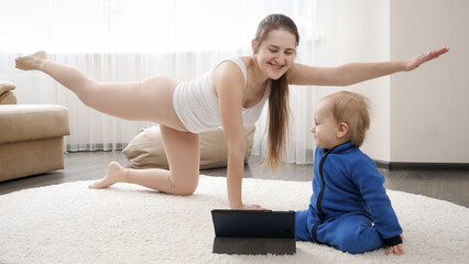Happy smiling mother with baby boy doing fitness by watching video on tablet computer. Family...