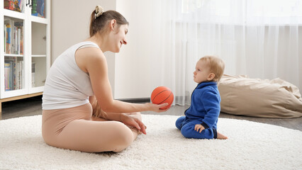 Young mother playing ball with her little baby son on soft carpet in living room. Family healthcare, active lifestyle, parenting and child development