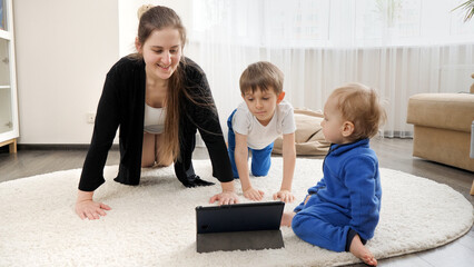 Young mother with two sons doing pushups with her little sons during online fitness lesson. Family healthcare, active lifestyle, parenting and child development